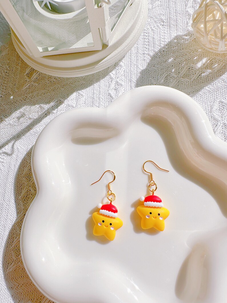 May include: A pair of gold-colored hook earrings. Each earring features a yellow star-shaped charm wearing a red and white Santa hat. The earrings are displayed on a white, wavy-edged plate.