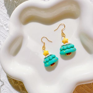 May include: A pair of Christmas tree earrings. Each earring features a teal tree with white frosting, a yellow star, and a small brown base. The earrings have gold-colored hooks and are displayed on a white, scalloped plate.