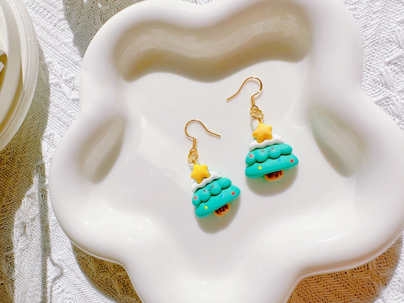 May include: A pair of Christmas tree earrings. Each earring features a teal tree with white frosting, a yellow star, and a small brown base. The earrings have gold-colored hooks and are displayed on a white, scalloped plate.