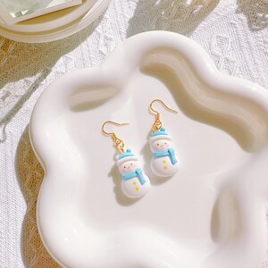 May include: A pair of snowman earrings with gold-colored hooks. Each earring features a white snowman with a blue hat and scarf, and yellow button details. The earrings are displayed on a white, scalloped-edge dish.