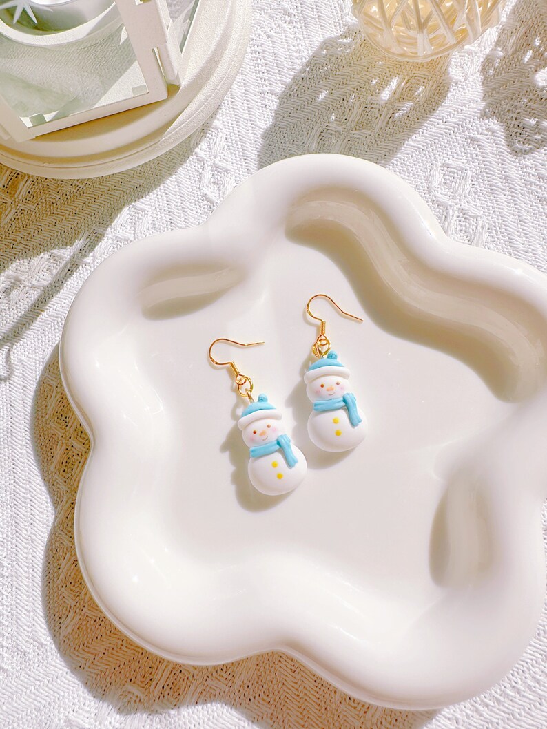 May include: A pair of snowman earrings with gold-colored hooks. Each earring features a white snowman with a blue hat and scarf, and yellow button details. The earrings are displayed on a white, scalloped-edge dish.