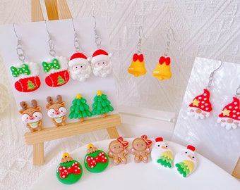 May include: A collection of Christmas-themed earrings and ornaments. The earrings feature designs like Santa Claus, Christmas stockings, bells, and gnomes. Ornaments include a green bauble with a red bow, gingerbread people, and a snowman. The earrings are displayed on white cards.