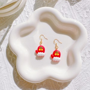 May include: A pair of festive earrings featuring a red mitten design with white trim and gold-colored hooks. The mittens have yellow accents and are displayed on a white, cloud-shaped dish. The earrings are a charming holiday accessory.