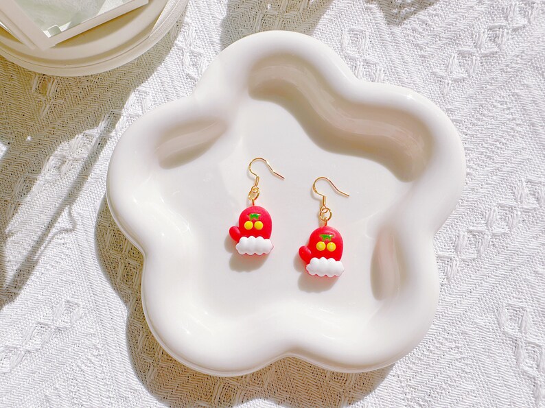 May include: A pair of festive earrings featuring a red mitten design with white trim and gold-colored hooks. The mittens have yellow accents and are displayed on a white, cloud-shaped dish. The earrings are a charming holiday accessory.