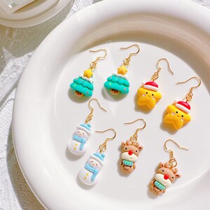 May include: A collection of Christmas-themed dangle earrings. The earrings feature designs of Christmas trees, snowmen, stars wearing Santa hats, and reindeer. Each earring has a gold-colored hook.