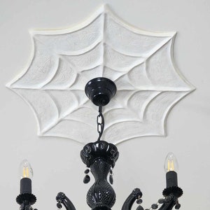 Puede incluir: Una araña de cristal negra con gotas de cristal cuelga de un techo blanco con un diseño de telaraña. La araña tiene cuatro brazos y tres bombillas.