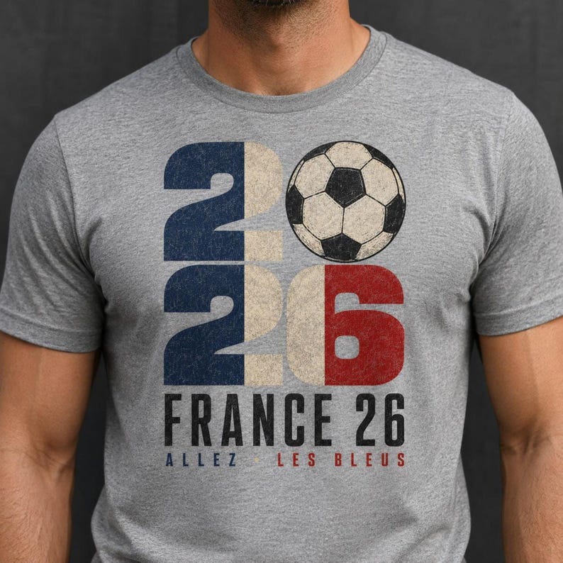 May include: A heather grey crew neck t-shirt featuring a large graphic with the number 2026, where the zero is replaced by a black and white football. Below the graphic, the text reads FRANCE 26 and ALLEZ LES BLEUS in black and red lettering.