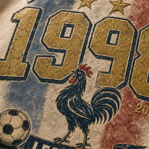 Puede incluir: Camiseta beige con un dise&ntilde;o desgastado que muestra el texto "FRANCE SOCCER 1998" en dorado y azul. Un gallo azul, un bal&oacute;n de f&uacute;tbol y la frase "LES BLEUS" tambi&eacute;n est&aacute;n presentes.