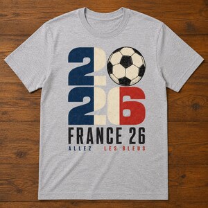 May include: A heather grey crew neck t-shirt featuring a large graphic with the number 2026, where the zero is replaced by a black and white football. The text reads FRANCE 26, ALLEZ, and LES BLEUS in blue, black, and red lettering.