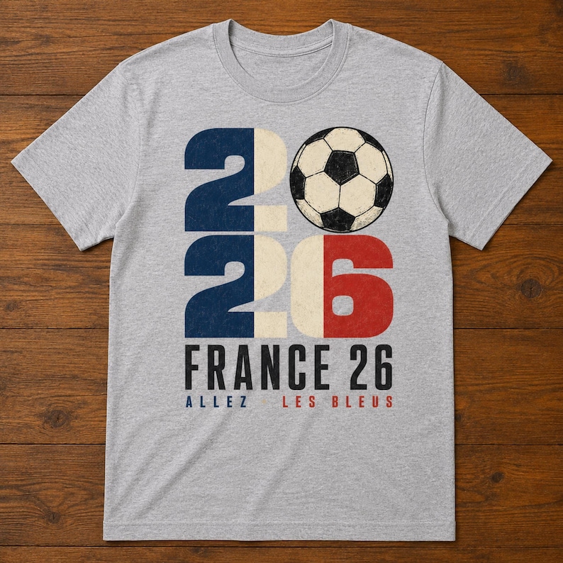 May include: A heather grey crew neck t-shirt featuring a large graphic with the number 2026, where the zero is replaced by a black and white football. The text reads FRANCE 26, ALLEZ, and LES BLEUS in blue, black, and red lettering.