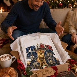 Puede incluir: Camiseta blanca con un dise&ntilde;o gr&aacute;fico que presenta "FRANCE WORLD CUP 26" en azul y rojo, un trofeo dorado e ilustraciones de monumentos franceses. La camiseta se presenta en una caja de cart&oacute;n con papel de seda, junto a una taza de chocolate caliente y cruasanes.