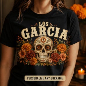 May include: A black short-sleeved t-shirt featuring a central graphic of a cream-coloured sugar skull surrounded by orange, red, and yellow flowers. The text "LOS GARCIA" is printed in a large, distressed beige font above the skull design.