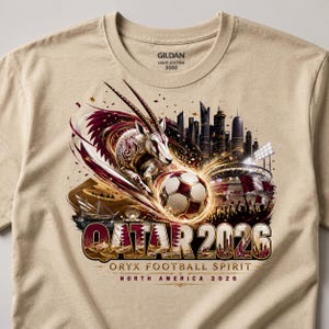 May include: A tan Gildan t-shirt featuring a graphic design for the Qatar 2026 World Cup. The design includes an oryx, a soccer ball, a stadium, and the text "QATAR 2026 ORYX FOOTBALL SPIRIT NORTH AMERICA 2026."