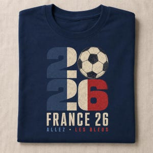 May include: A navy blue t-shirt featuring a graphic with the number 2026, where the zero is replaced by a football. The design incorporates the colours of the French flag and includes the text FRANCE 26 and ALLEZ LES BLEUS in a distressed font.
