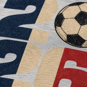 May include: A light grey fabric featuring a graphic print with large, bold letters in navy blue, tan, and red. The design includes a detailed illustration of a black and white football positioned near the top right corner of the printed text.