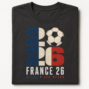 May include: A charcoal grey t-shirt featuring a graphic with the number 2026, where the zero is replaced by a black and white football. The design incorporates blue, white, and red colours, with the text FRANCE 26 and ALLEZ LES BLEUS below.