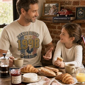 Puede incluir: Un hombre y una ni&ntilde;a est&aacute;n sentados a una mesa de madera, desayunando. El hombre lleva una camiseta beige con el texto "France Soccer 1998" y un gallo. La mesa tiene cruasanes, pan, queso, mermelada, caf&eacute; y zumo de naranja.