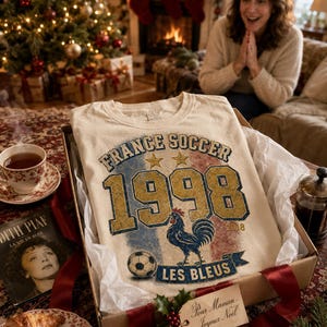 Puede incluir: Una camiseta color crema en una caja de regalo con el texto "FRANCE SOCCER 1998 LES BLEUS". La camiseta presenta un bal&oacute;n de f&uacute;tbol y un gallo. Una mujer est&aacute; en el fondo y se ve un &aacute;rbol de Navidad.