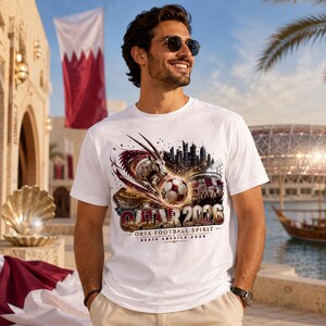 May include: White t-shirt with a graphic featuring a soccer ball, an oryx, and the text "QATAR 2026". The design includes a cityscape and stadium imagery, with the words "Oryx Football Spirit - North America 2026" below.