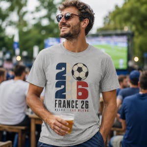 May include: A heather grey short-sleeve t-shirt featuring a graphic with the number 2026, a football, and the text FRANCE 26 ALLEZ LES BLEUS. The design uses navy blue, red, and white colours with a distressed texture on the front of the shirt.