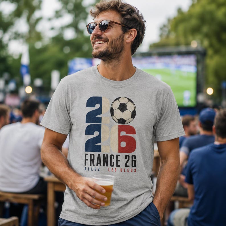 May include: A heather grey short-sleeve t-shirt featuring a graphic with the number 2026, a football, and the text FRANCE 26 ALLEZ LES BLEUS. The design uses navy blue, red, and white colours with a distressed texture on the front of the shirt.