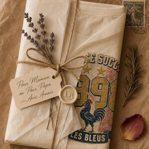 Puede incluir: Un regalo envuelto en papel marr&oacute;n, atado con hilo y adornado con ramitas de lavanda. El regalo incluye una camiseta con un gr&aacute;fico de gallo y el texto "SE SOCC 1991 LES BLEUS". Una etiqueta dice "Pour Maman ou Pour Papa - Avec Amour".