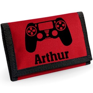 Puede incluir: Cartera roja con ribete negro, con un diseño de mando de juego negro y el nombre "Arthur" impreso debajo. La cartera es rectangular y parece estar hecha de tela duradera.