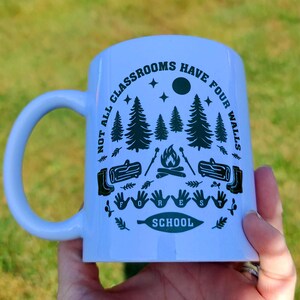 White mug with green woodland design showing trees, stars, moon, campfire, and boots, printed with “Not all classrooms have four walls – Forest School,” photographed in natural light on grass.