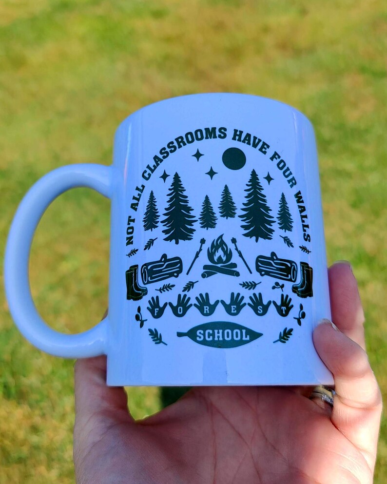 White mug with green woodland design showing trees, stars, moon, campfire, and boots, printed with “Not all classrooms have four walls – Forest School,” photographed in natural light on grass.