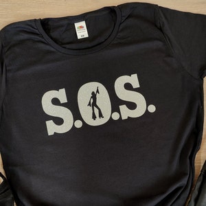 Puede incluir: Camiseta negra con un diseño de purpurina plateada que dice "S.O.S." con una silueta de una mujer bailando en la "O".