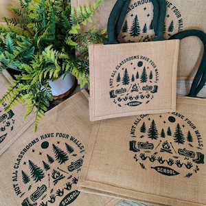 A group of jute tote bags with teal rope handles arranged on a light surface beside a green fern plant. Each bag features a dark green outdoor-themed print showing pine trees, logs, a campfire, and handprints, with the words “Not all classrooms have four walls – School.” The natural textures and earthy tones highlight an eco-friendly, outdoor learning theme.
