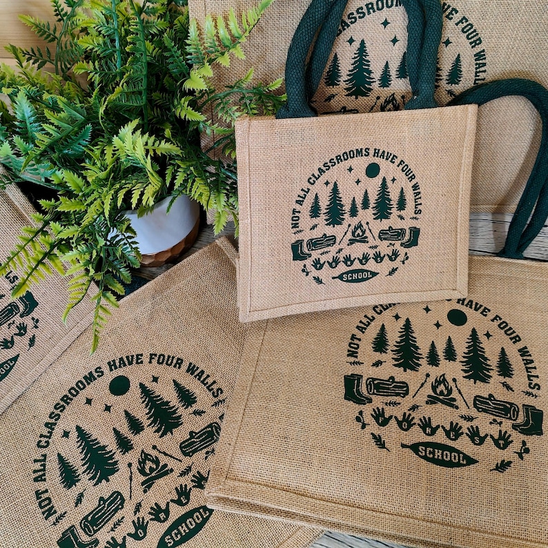 A group of jute tote bags with teal rope handles arranged on a light surface beside a green fern plant. Each bag features a dark green outdoor-themed print showing pine trees, logs, a campfire, and handprints, with the words “Not all classrooms have four walls – School.” The natural textures and earthy tones highlight an eco-friendly, outdoor learning theme.