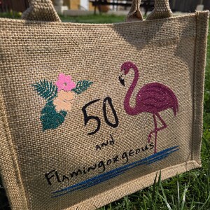 A natural jute tote bag lying on grass, featuring a glittery pink flamingo standing on one leg, tropical flowers in pink and peach with green leaves, and black and blue text reading “Flamingo Bay Bags 50 and.” The design has a bright, summery, eco-friendly look with a textured burlap surface.