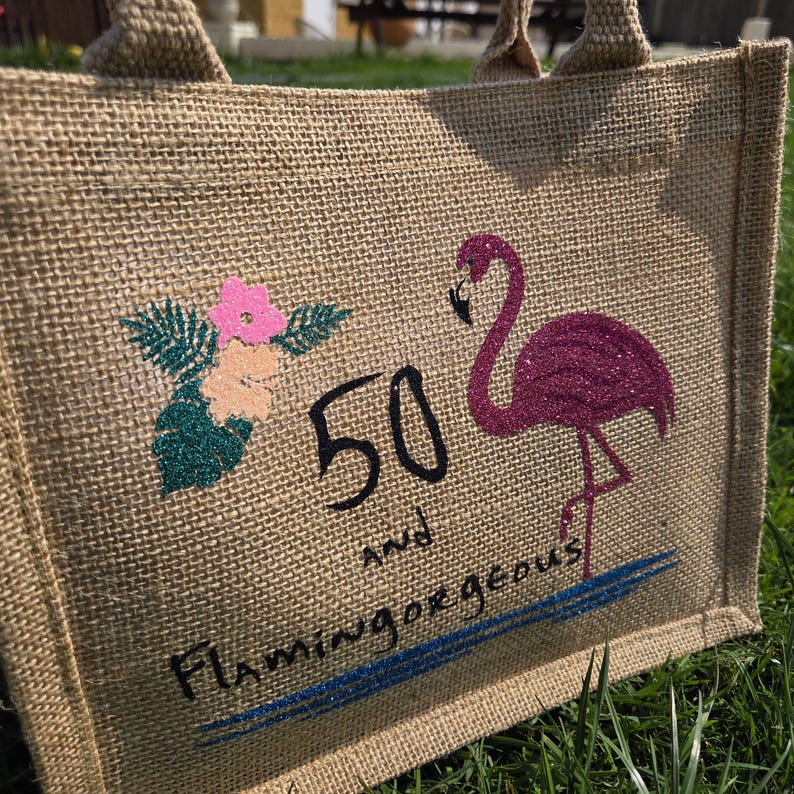 A natural jute tote bag lying on grass, featuring a glittery pink flamingo standing on one leg, tropical flowers in pink and peach with green leaves, and black and blue text reading “Flamingo Bay Bags 50 and.” The design has a bright, summery, eco-friendly look with a textured burlap surface.