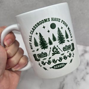 White ceramic mug with green forest‑school design featuring pine trees, campfire, logs, boots, and the phrase “Not all classrooms have four walls – Forest School,” held in a hand above a light wooden surface beside a woven basket and grey patterned plant pot.