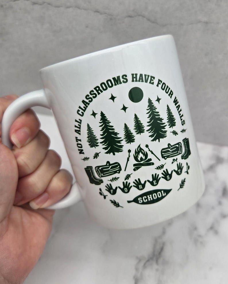 White ceramic mug with green forest‑school design featuring pine trees, campfire, logs, boots, and the phrase “Not all classrooms have four walls – Forest School,” held in a hand above a light wooden surface beside a woven basket and grey patterned plant pot.