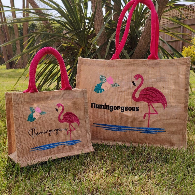 Two brown jute tote bags with pink flamingo designs and the text "Flamingorgeous". The larger bag has a pink flamingo standing on one leg in blue water. The smaller bag has a pink flamingo standing on one leg in blue water with a pink flower and green leaf design.