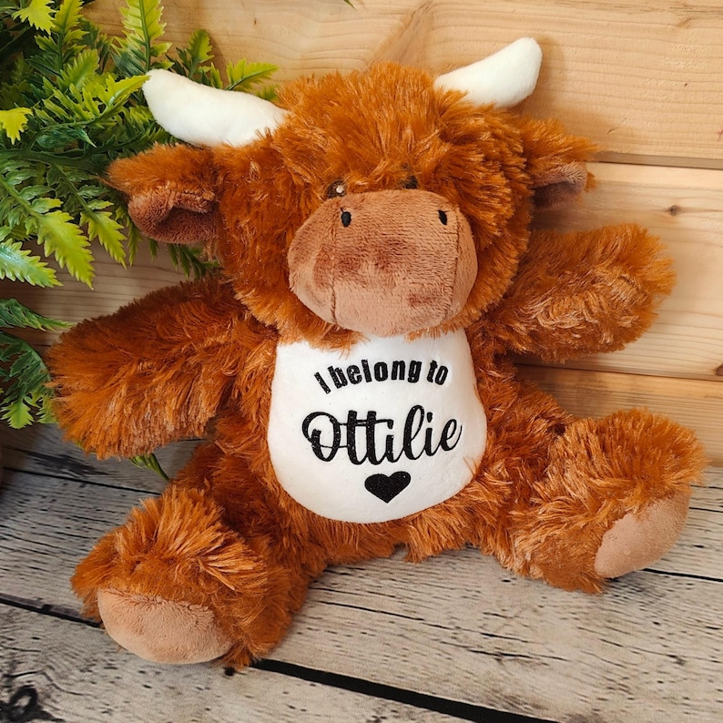 May include: Brown and white plush Highland cow stuffed animal with a white bib that says "I belong to Ottilie" with a black heart.