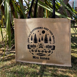 A Jute tote bag with black handles. Each bag features a circular design with trees, a campfire, and the text "NOT ALL CLASSROOMS HAVE FOUR WALLS SCHOOL". The bags are on grass.