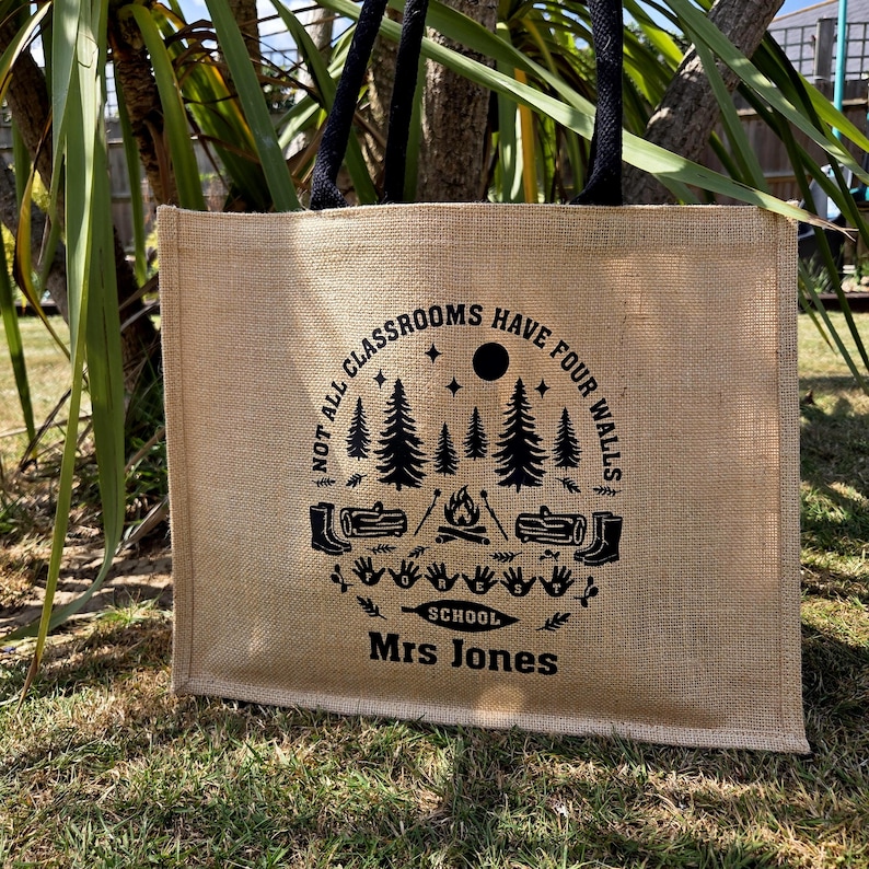 A Jute tote bag with black handles. Each bag features a circular design with trees, a campfire, and the text "NOT ALL CLASSROOMS HAVE FOUR WALLS SCHOOL". The bags are on grass.