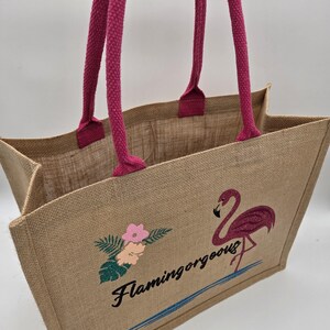 Natural colour jute bag with pink handle. Displays a pink flamingo using glitter htv, standing in a glitter blue lake, with flowers to one side, personalised with flamingorgeous in a black glitter font.