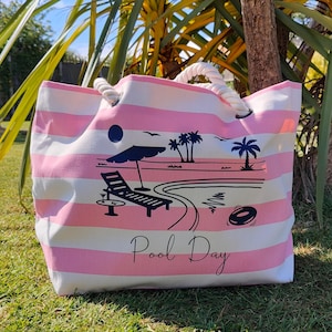 Puede incluir: Bolso tote a rayas rosas y blancas con asas de cuerda. El bolso presenta un gráfico de una escena de piscina con una tumbona, una sombrilla y las palabras "Pool Day".