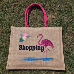 Natural colour jute bag with pink handle. Displays a pink flamingo using glitter htv, standing in a glitter blue lake, with flowers to one side, personalised with Shopping