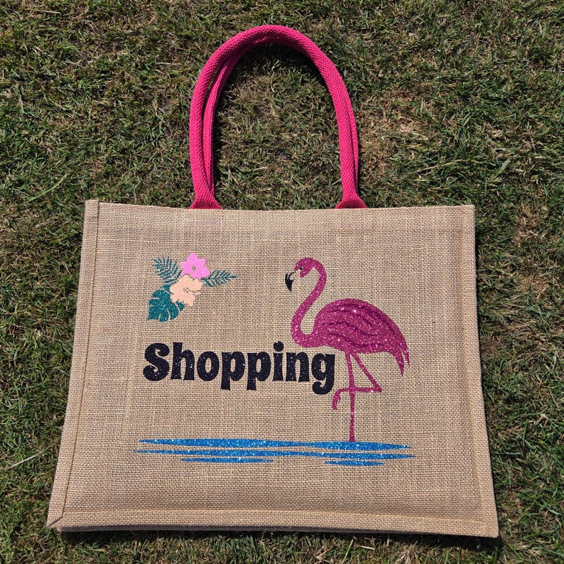 Natural colour jute bag with pink handle. Displays a pink flamingo using glitter htv, standing in a glitter blue lake, with flowers to one side, personalised with Shopping