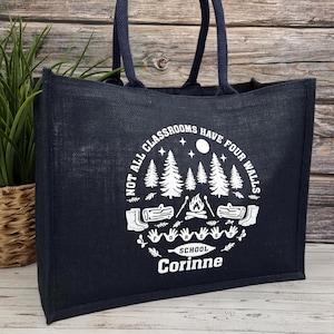 photo of a navy jute bag placed on a wooden side. The bag features a  white print showing trees, logs, a campfire, and handprints with the words “Not all classrooms have four walls – School.”