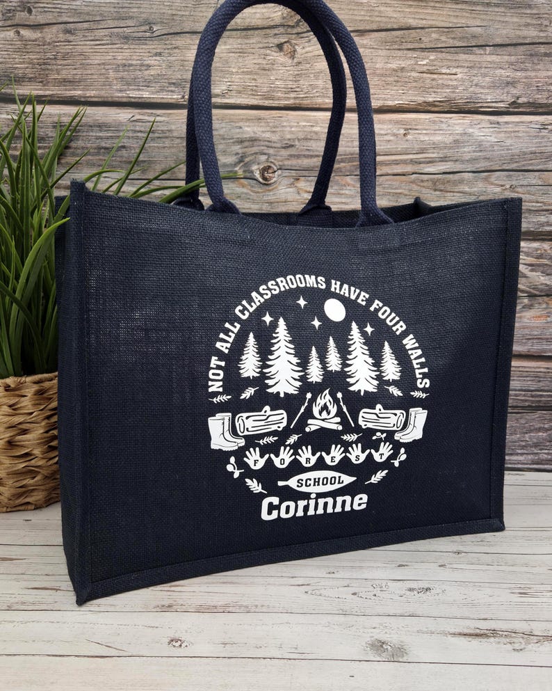 photo of a navy jute bag placed on a wooden side. The bag features a  white print showing trees, logs, a campfire, and handprints with the words “Not all classrooms have four walls – School.”