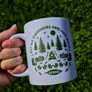 White ceramic mug held outdoors against green grass, featuring a forest‑themed design with pine trees, campfire, logs, boots, and the phrase “Not all classrooms have four walls – Forest School.”