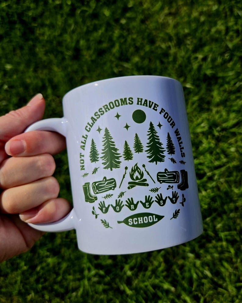 White ceramic mug held outdoors against green grass, featuring a forest‑themed design with pine trees, campfire, logs, boots, and the phrase “Not all classrooms have four walls – Forest School.”