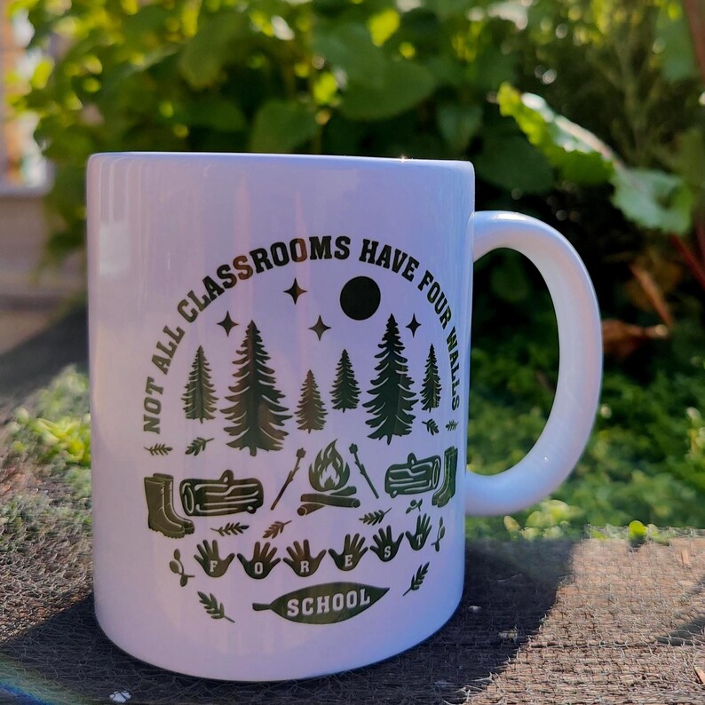 Forest School mug on a wooden surface with leafy plants in the background, showing green outdoor‑learning design with trees, campfire, and boots.