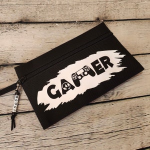 Può includere: Astuccio nero con chiusura a zip e cinturino da polso. La parola "GAMER" è stampata in bianco, insieme a una grafica di un controller di gioco. Un piccolo ciondolo di perline con il nome "JACOB" pende dal cinturino.